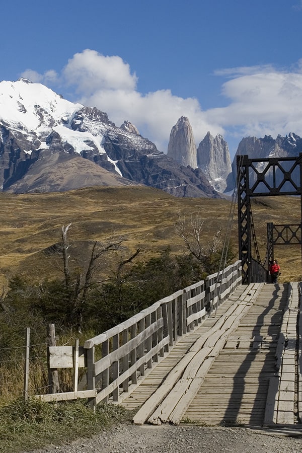 A visitor crossing a bridge in Torres del Paine National Park, with a couple of the peaks visible.
