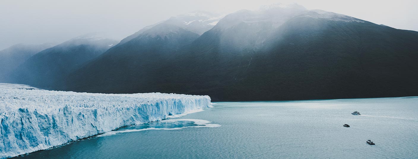 Boats in the water near a massive glacier in Patagonia, with snowcapped mountains overhead.