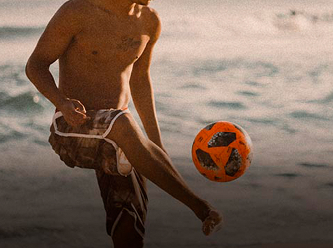 A Brazilian man kicking a soccer ball into the air with his right foot on a sandy beach in Brazil