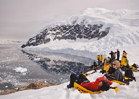 A group of hikers sitting on a snowbank and resting while looking at the icy sea in Antarctica.