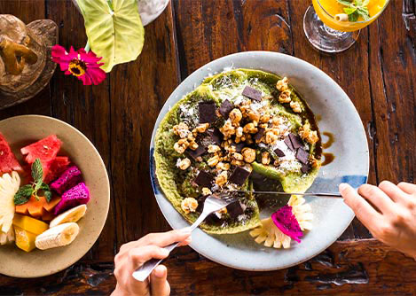 Plates of colorful Brazilian food on a wooden table, with a diner cutting into one of the dishes.